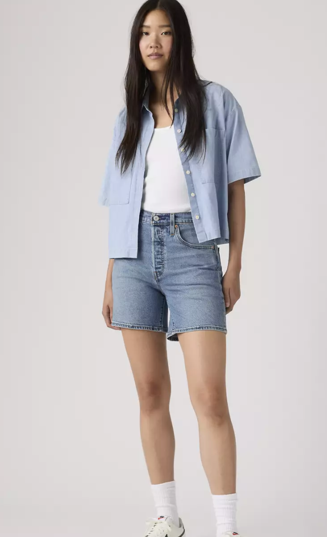 Person wearing a light blue shirt, white top, and denim shorts on a plain background
