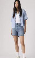 Person wearing a light blue shirt, white top, and denim shorts on a plain background