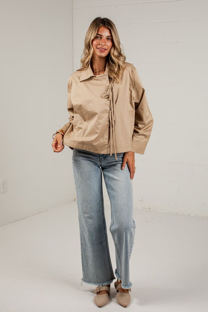 A woman modeling a light tan utility jacket with front tie details, paired with wide-leg light blue jeans and beige pointed-toe flats, standing against a white brick wall.