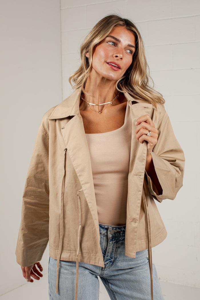 A woman wearing a light tan utility jacket over a beige tank top and blue jeans, standing against a white brick wall.