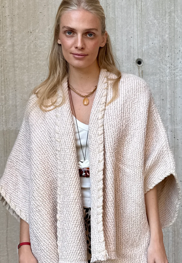 Woman wearing a beige knitted shawl against a concrete wall.