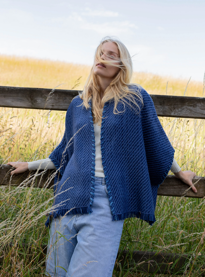 Woman wearing a blue knitted poncho standing in a field with a wooden fence.