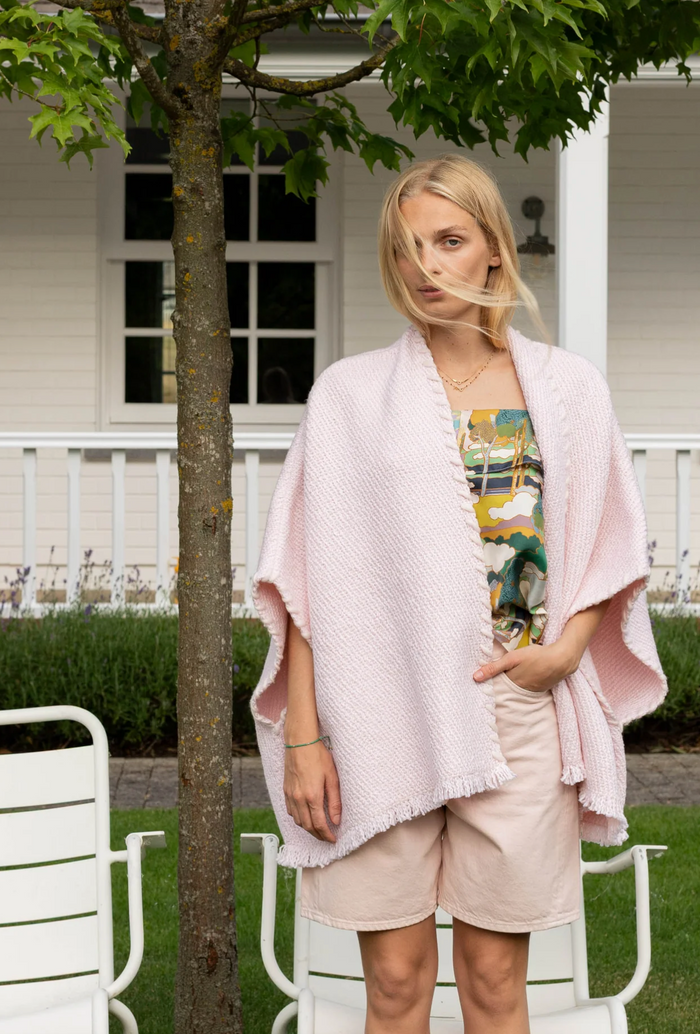 Woman wearing a pink shawl in front of a white house with greenery.