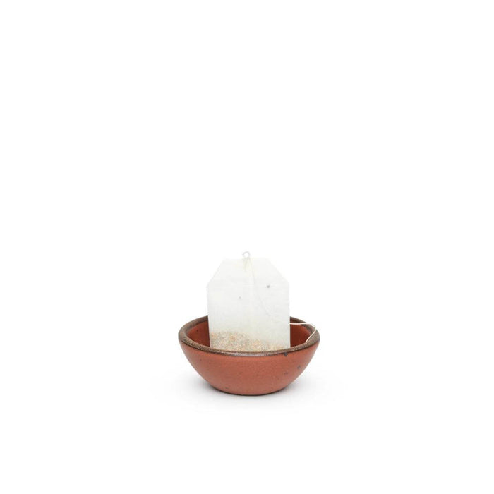 A tiny rounded ceramic bowl in a cool burnt terracotta color featuring iron speckles and an unglazed rim, with a teabag