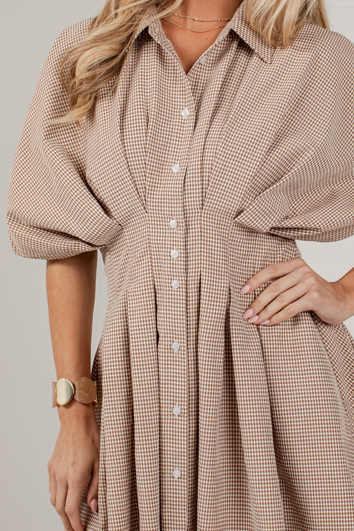 A close-up of a woman wearing a beige and brown gingham button-up shirt dress with voluminous elbow-length sleeves and a pleated skirt, photographed against a plain light gray studio background.
