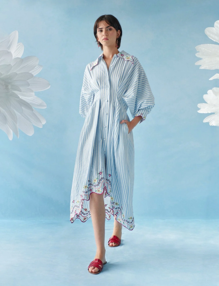 Woman wearing a long, light blue and white striped dress with floral embroidery on a light blue background.