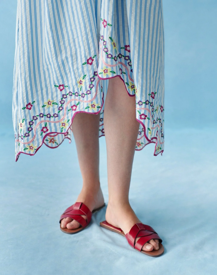 Person wearing a striped dress with floral embroidery and red sandals on a light blue background