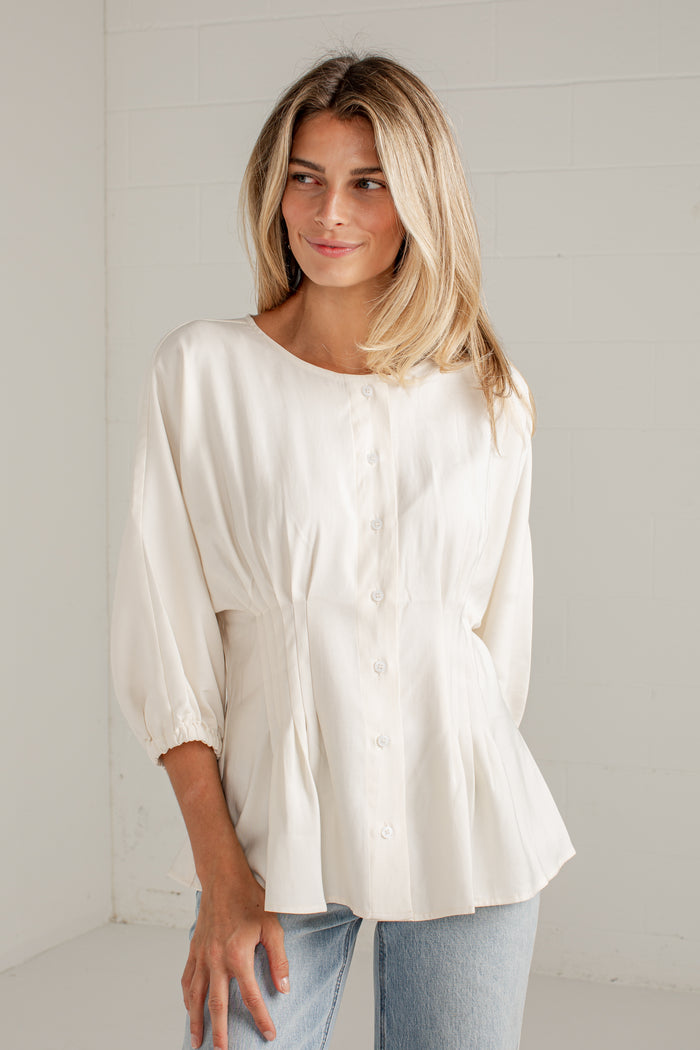A woman wearing a cream-colored, long-sleeved button-up blouse with a gathered peplum waist, paired with light blue jeans, standing against a white brick wall.