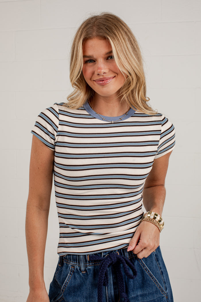 A woman wearing a short-sleeve crewneck t-shirt with navy blue and cream horizontal stripes, paired with blue denim jeans, standing against a white brick wall.