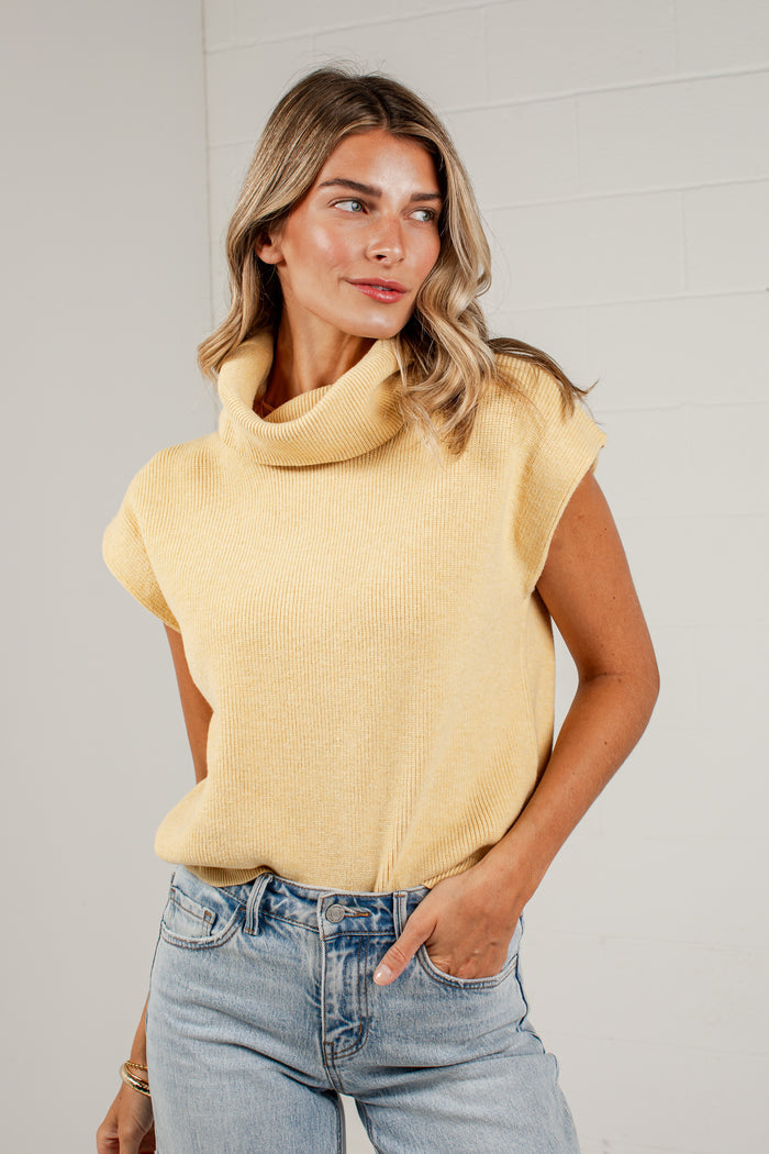 A woman wearing a pale yellow ribbed knit turtleneck sweater with cap sleeves and light-wash blue jeans, standing against a white brick wall.