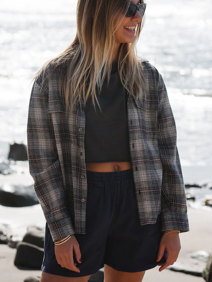 Woman wearing a plaid shirt over a black top and navy shorts by a waterfront.
