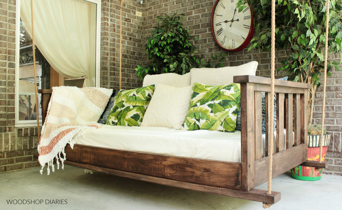 Finished DIY porch swing bed with white cushion, tassel throw blanket, and tropical leaf pillows on brick porch