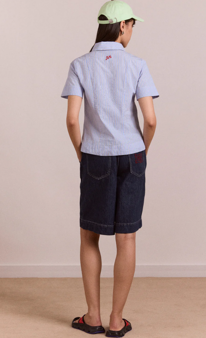 Person wearing a light blue checkered shirt and dark shorts with a green cap, standing against a plain background.