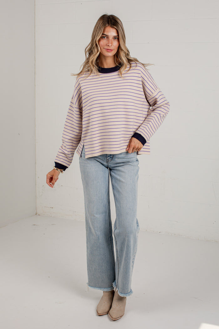 A woman modeling a long-sleeve striped knit top with navy blue trim, paired with light-wash wide-leg jeans and beige ankle boots, standing in a bright white studio corner.
