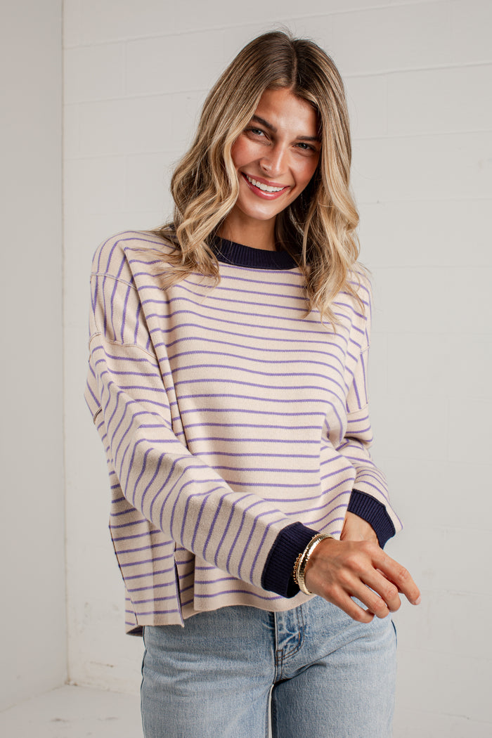 A woman wearing a lavender and navy blue horizontally striped long-sleeve knit sweater with solid navy ribbed cuffs and collar, paired with light-wash blue jeans, standing against a white paneled wall.