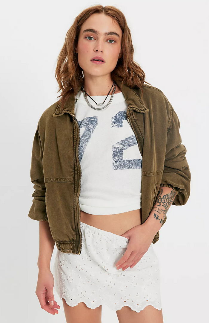 Woman wearing an olive green jacket over a white graphic t-shirt and ivory lace skort on a white background