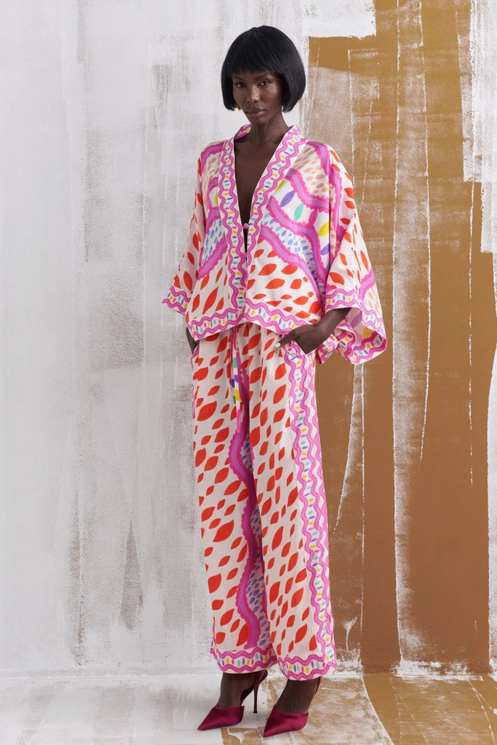 A model wearing a matching two-piece lounge set with a bold pink, orange, and white patterned print, standing against a textured beige and white wall.
