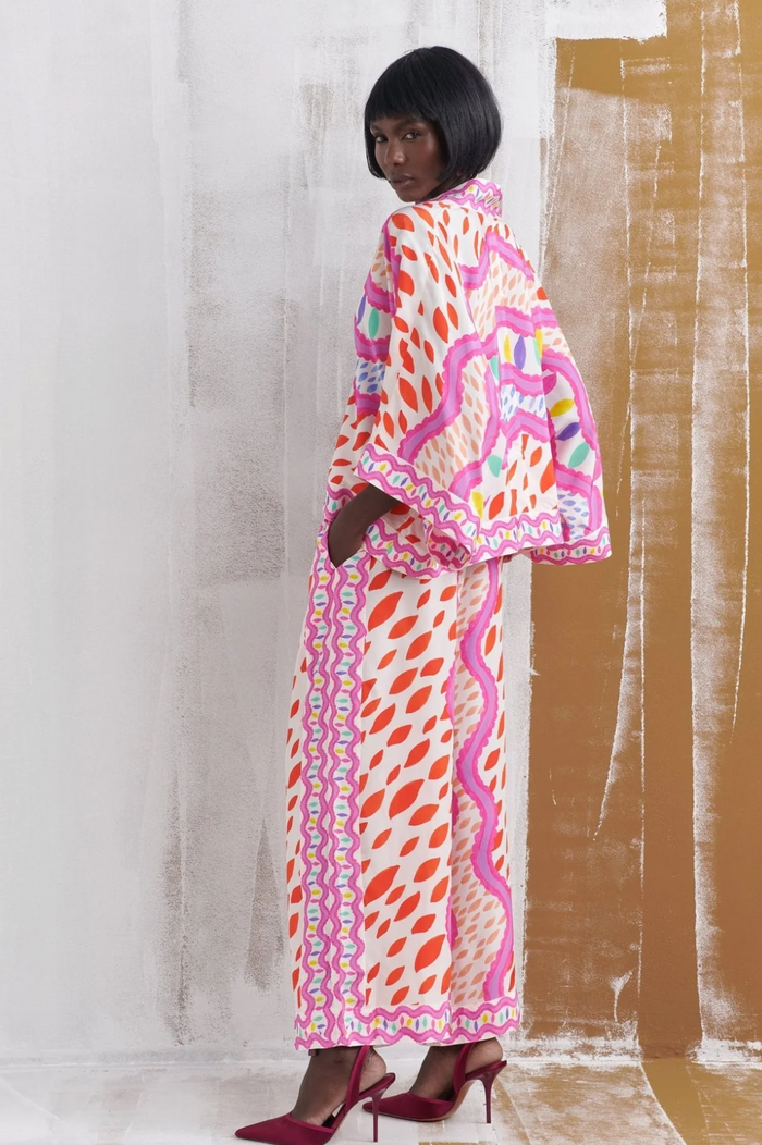 A model wearing a long white kaftan dress with a vibrant pink, orange, and green abstract pattern, paired with dark red heels, standing against a textured studio backdrop.