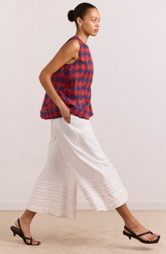 Woman wearing a red and blue patterned top and white pants against a plain background