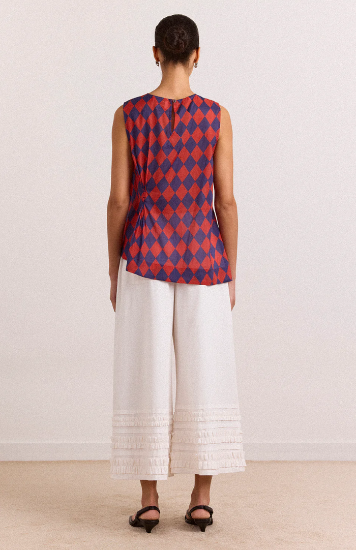 Woman wearing a red and blue patterned sleeveless top with white pants on a plain background