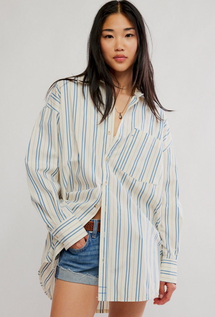 A woman modeling an oversized long-sleeve button-up shirt with vertical blue and green stripes on a cream background, paired with denim shorts against a light gray studio background.