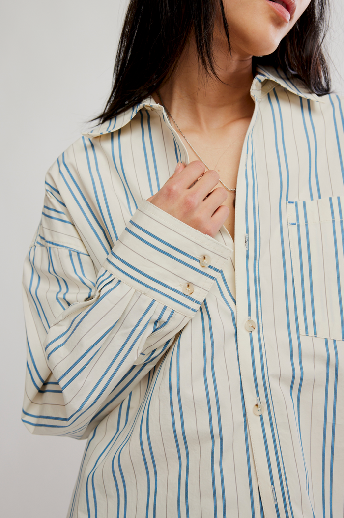 A close-up of a model wearing an oversized ivory-colored button-up shirt with vertical blue stripes. The model is shown from the chest up, with her hand resting on the front of the shirt.