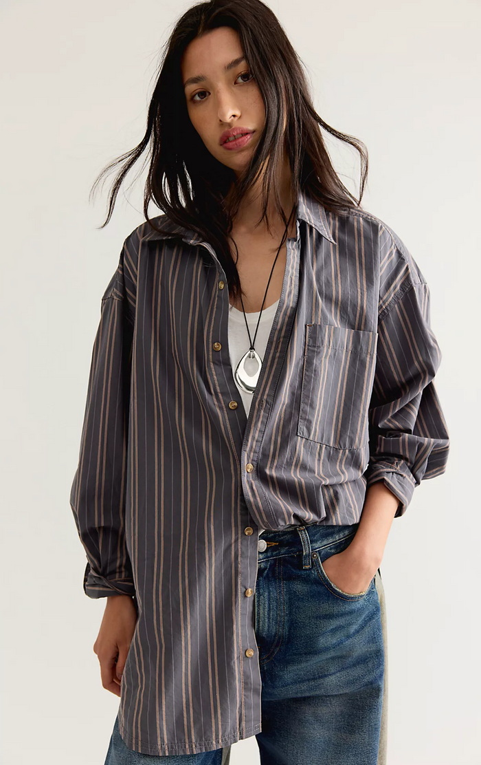 A woman wearing an oversized button-up shirt with vertical stripes in gray, brown, and beige, layered over a white top and paired with blue jeans.