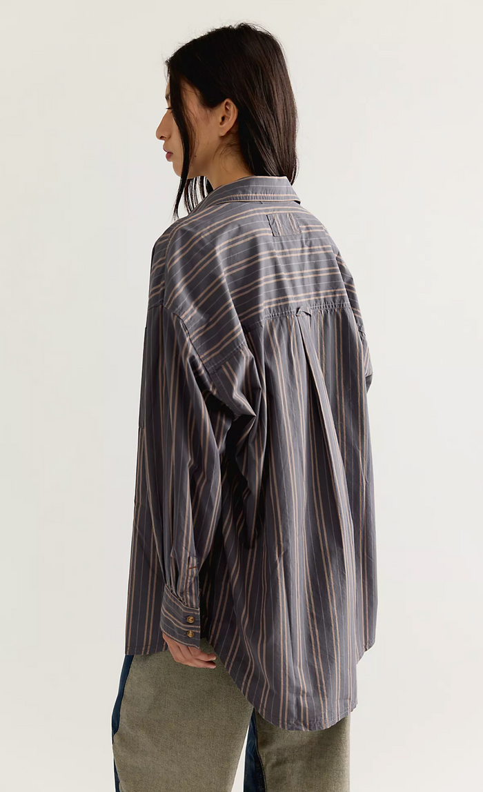 A model shown from the back wearing an oversized, long-sleeve striped button-up shirt in shades of gray, blue, and white, paired with olive green pants against a plain white studio background.