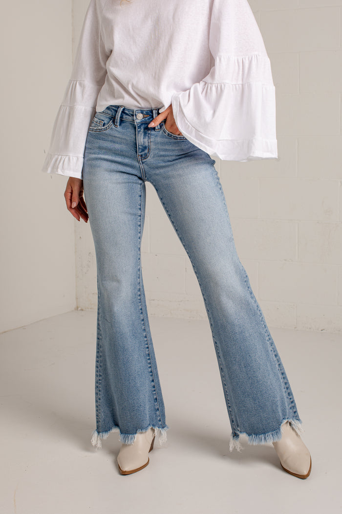A person wearing light-wash blue flare jeans with frayed hems, paired with a white tiered blouse and beige pointed-toe boots, standing against a light neutral wall.