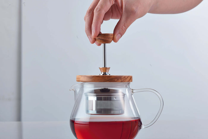 Hand pulling up metal tea strainer of glass teapot with wooden lid filled with tea, on a light gray background