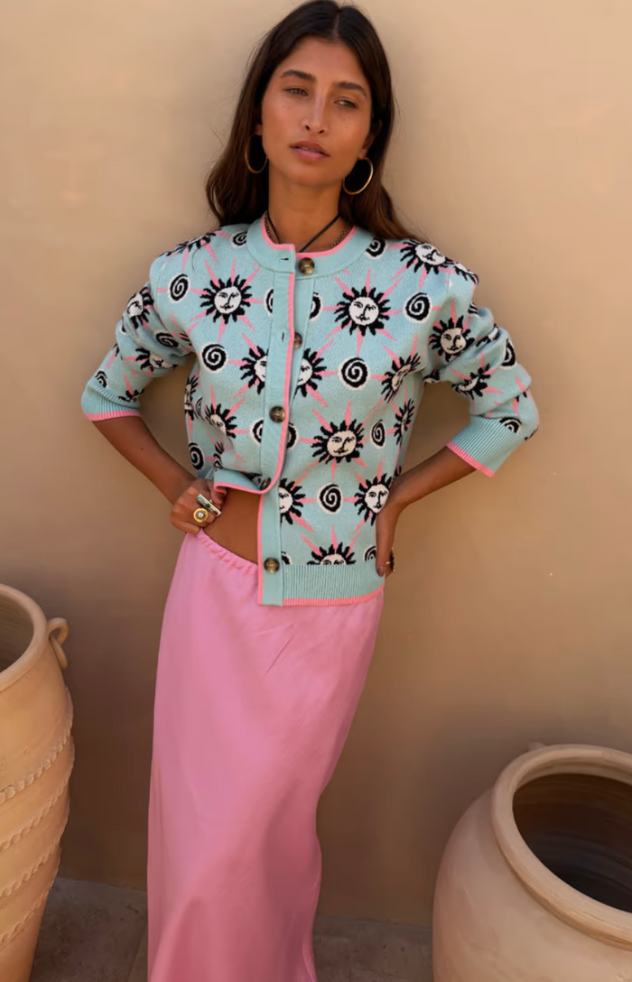 Woman wearing a patterned light blue jacket and pink pants against a beige wall.