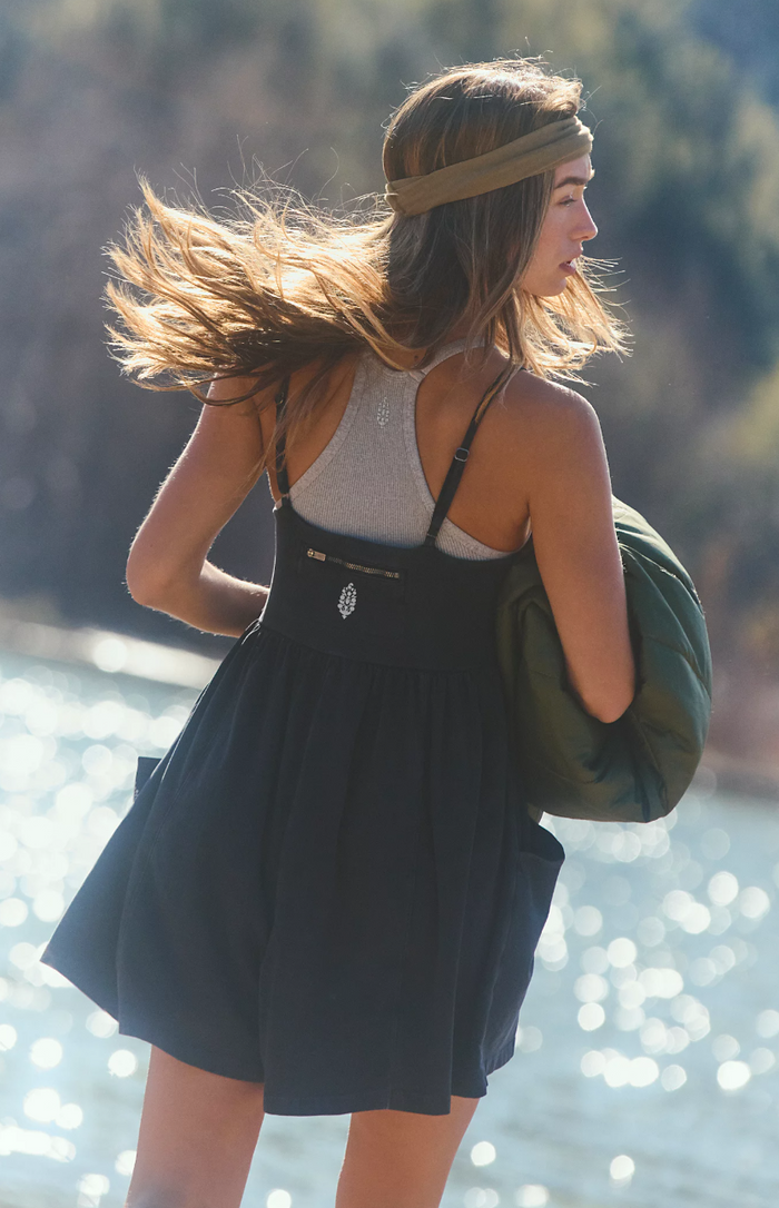 Woman with long hair and a green headband standing by a body of water, wearing a black dress and carrying a green bag.