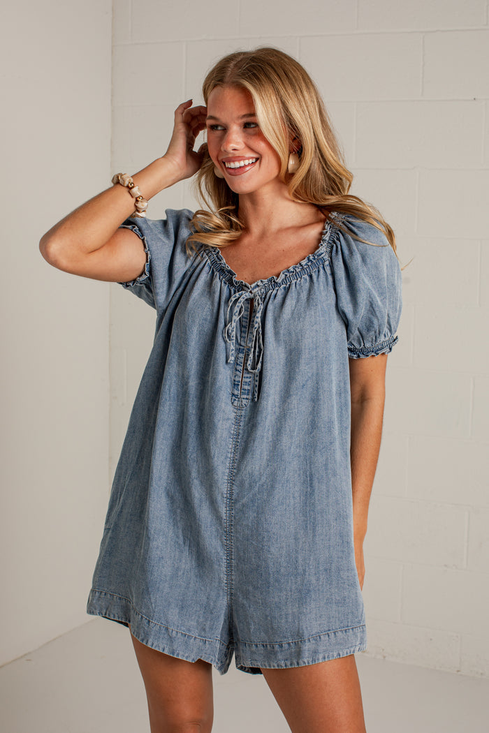 A woman modeling a light blue denim short romper with puff sleeves and a tie neckline, standing against a white brick wall.