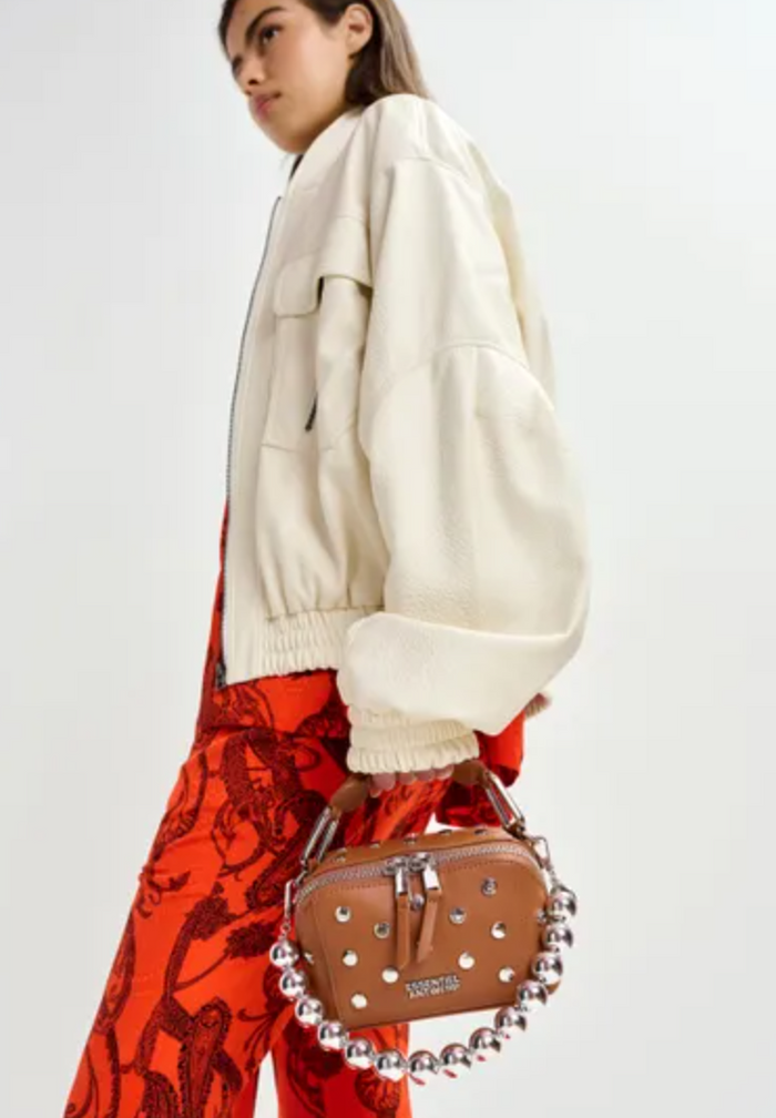 Person wearing a cream jacket and red patterned pants holding a brown handbag with silver studs on a white background