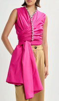 Woman wearing a pink sleeveless dress with a ruched front and beige pants on a white background