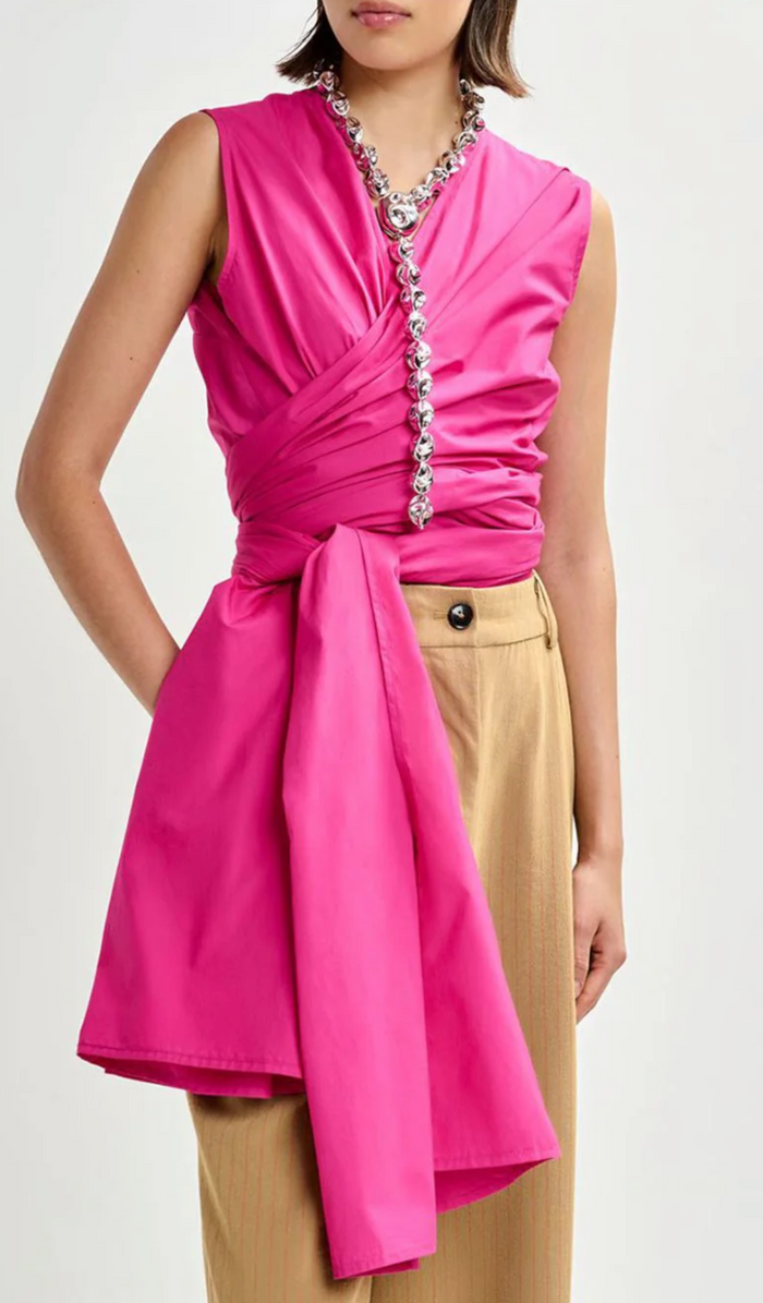 Woman wearing a pink sleeveless dress with a ruched front and beige pants on a white background