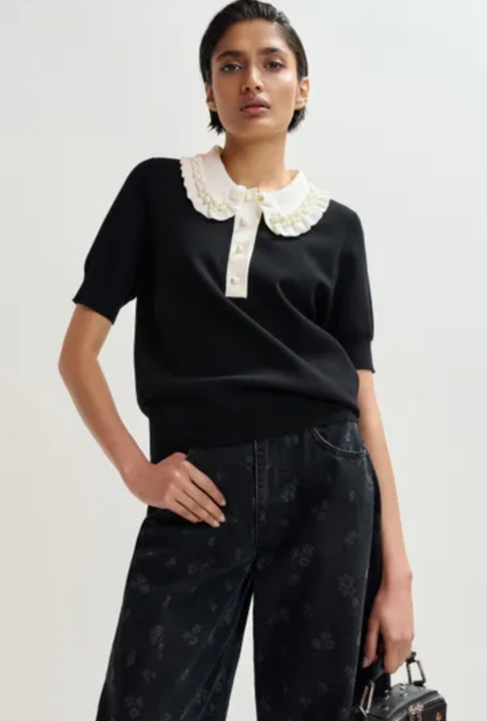 Woman wearing a black top with a white collar and dark pants on a plain background