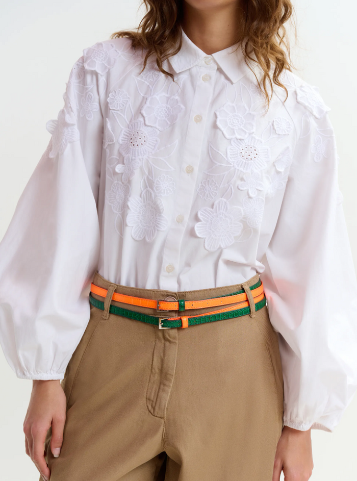 Person wearing a white blouse with floral embroidery and beige pants with an orange belt.