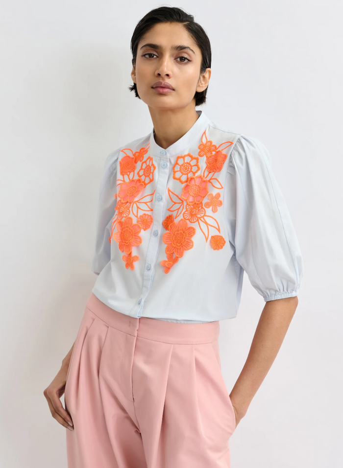 Woman wearing a light blue blouse with orange floral embroidery and pink pants against a white background