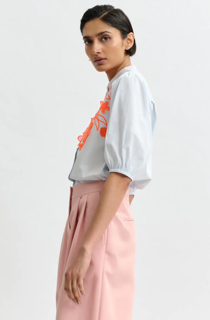 Woman wearing a white blouse with orange accents and pink pants on a light gray background