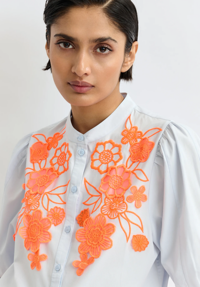 Woman wearing a white shirt with orange floral embroidery on a plain background
