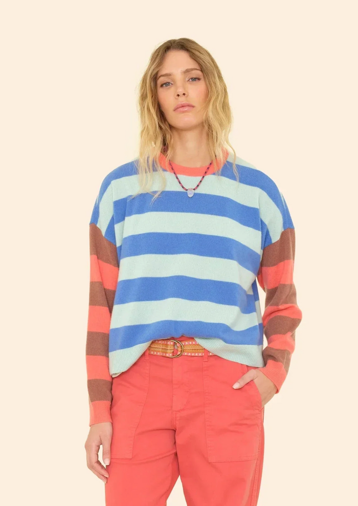 A woman modeling a long-sleeve crewneck sweater with horizontal stripes in light blue, medium blue, coral pink, and brown, paired with coral pants, against a solid cream background.