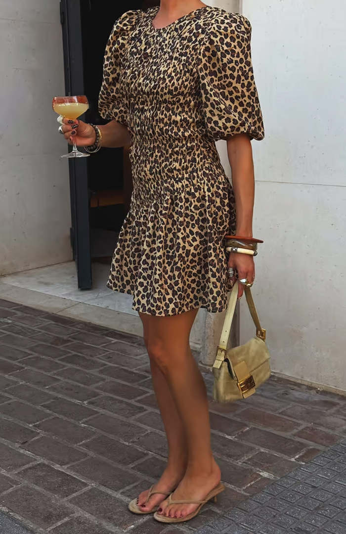 Person wearing a leopard print dress holding a drink and a handbag on a sidewalk.