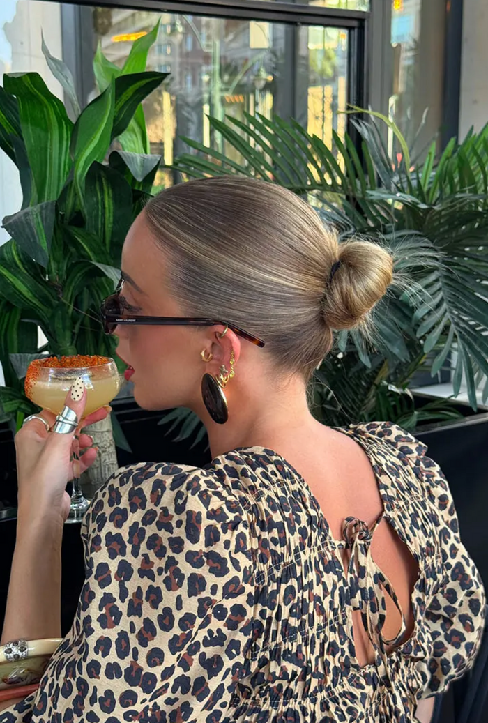 Woman in leopard print dress holding a drink in an indoor setting with plants.