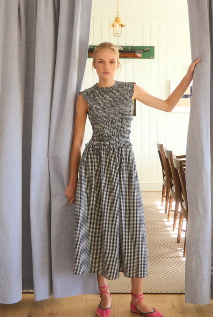 Woman in a checkered dress standing between two curtains in a room with a dining table.