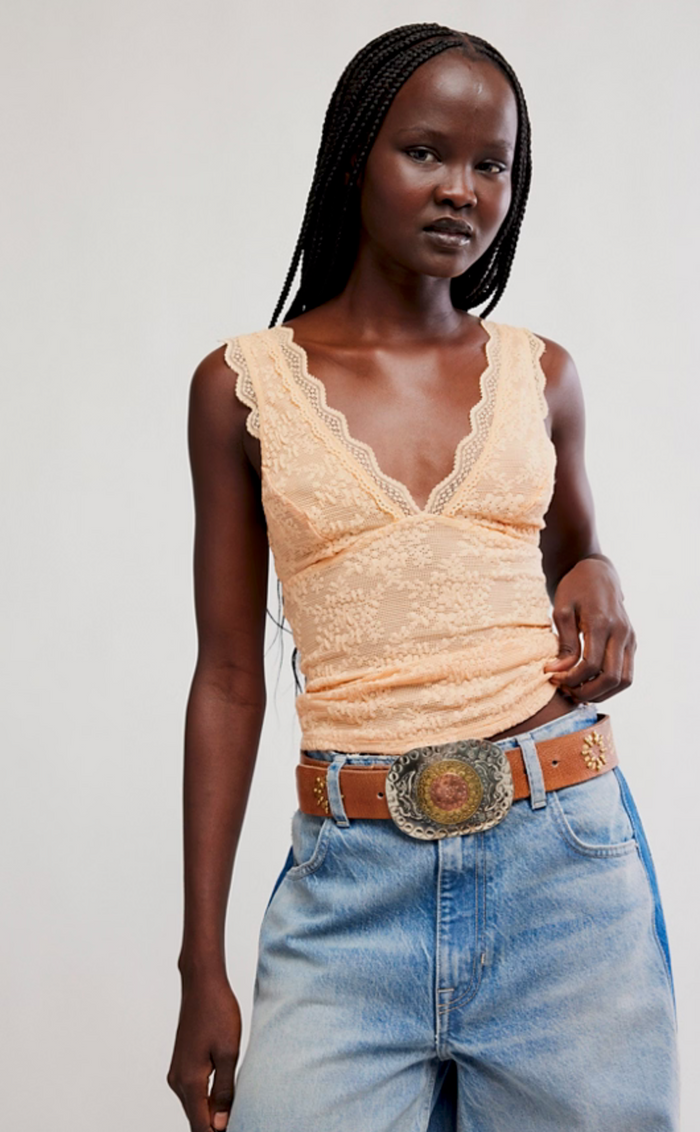 Woman wearing a peachy lace top and blue jeans with a large belt buckle on a plain background