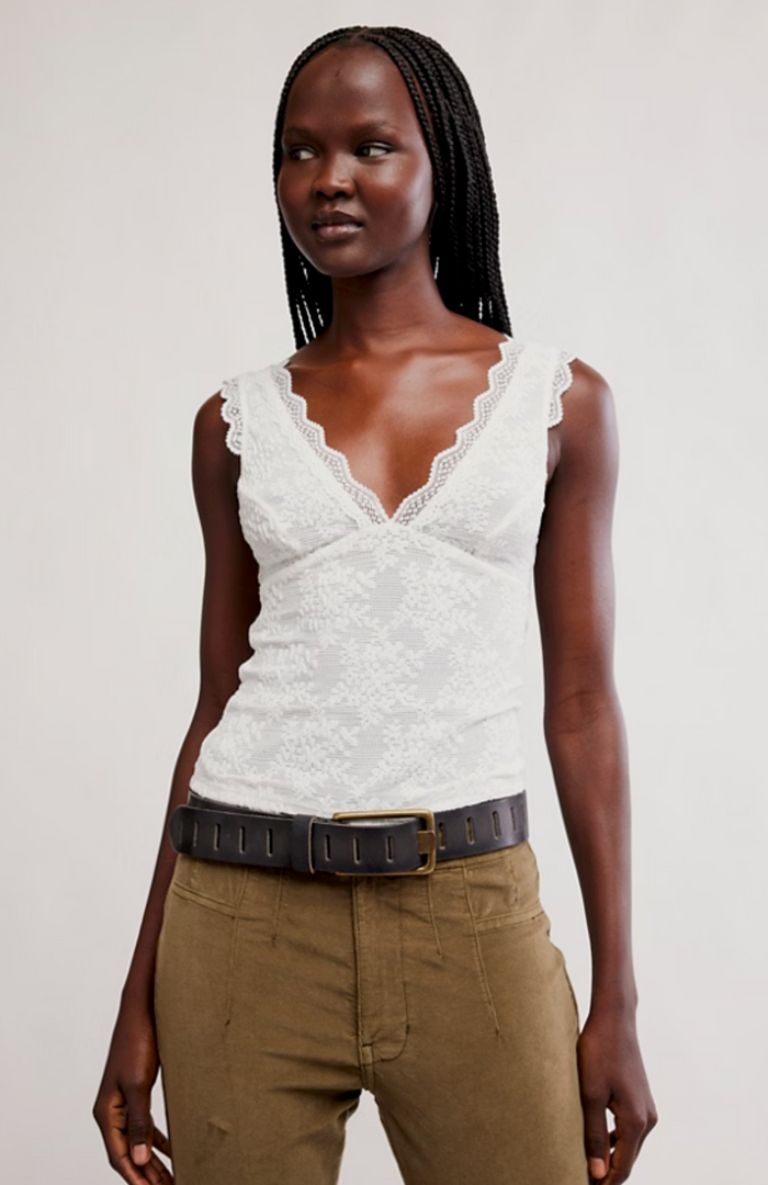 Woman wearing a white sleeveless top with lace details and brown pants on a plain background