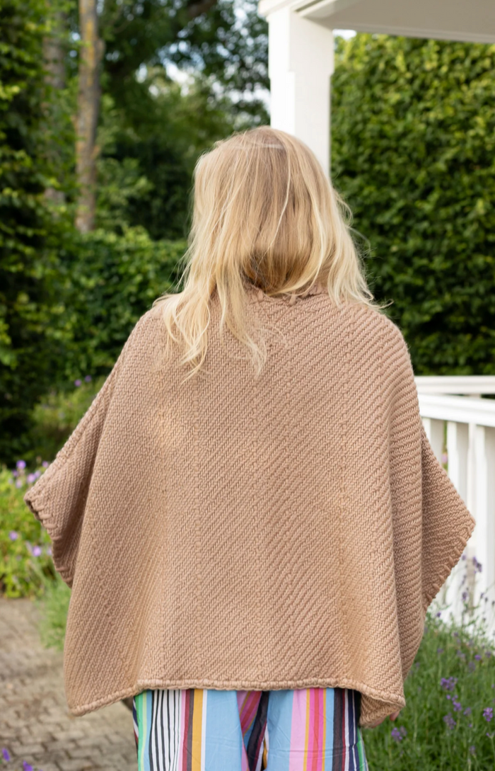Person wearing a beige poncho in an outdoor setting with greenery and a white structure.