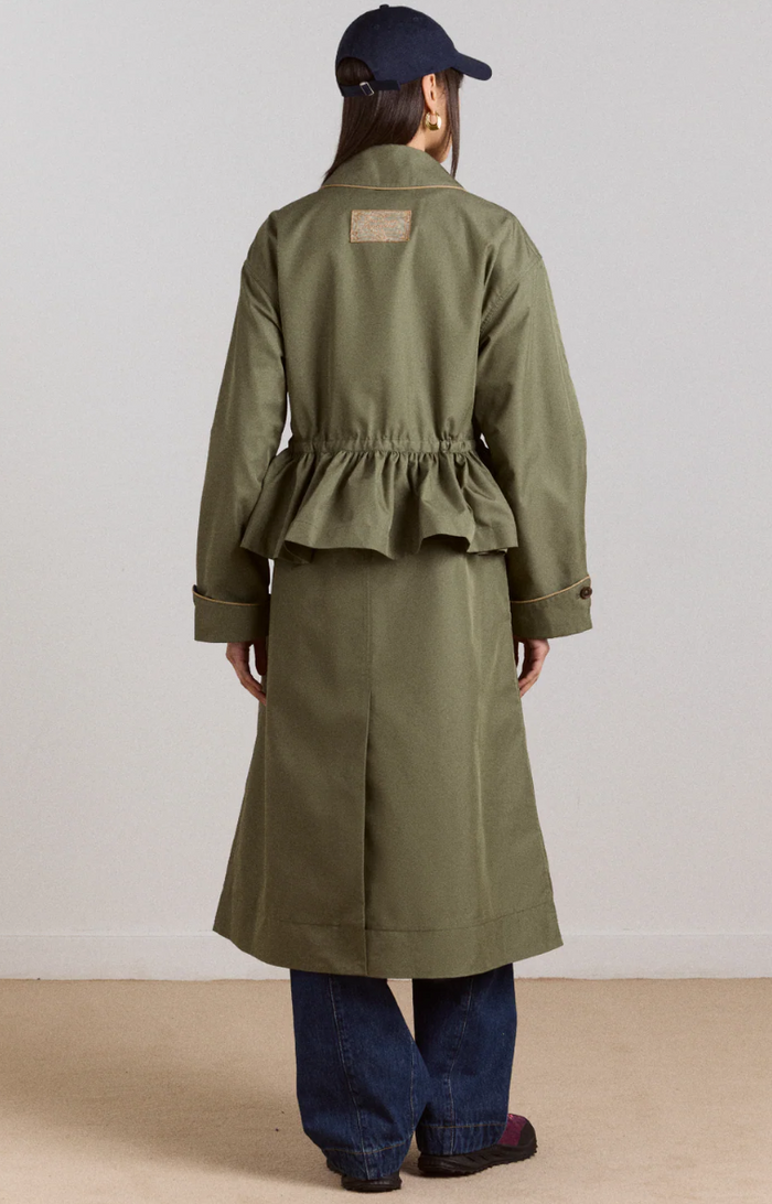 Person wearing a sage trench coat and dark cap on a plain background