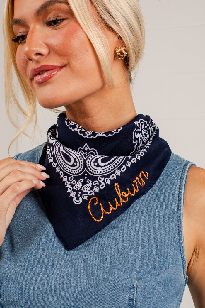 A close-up of a woman wearing a navy blue bandana with a white paisley pattern and the word 'Cubum' embroidered in orange on the front.